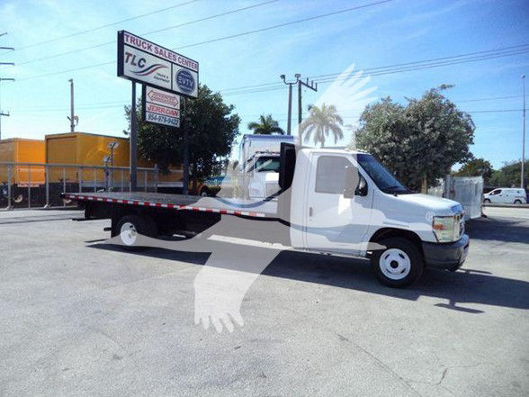Ford E-450 flatbed tow truck with long low bed, white cab, and black chassis. Visible signage includes a TLC Truck Sales Center sign in the background and a palm tree graphic watermark overlay on the image.