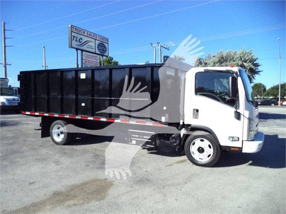 Isuzu NPRR dump truck in a used truck lot, white cab, black dump body, DOT wheels, stock number 18FTDUMPNRRFM visible in background.