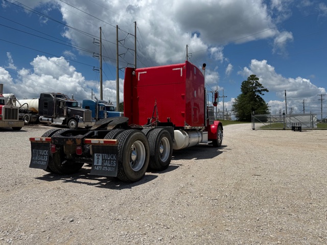 Peterbilt 379 semi truck red sleeper cab viewed from rear right side, multiple axles, gravel lot, blue sky with clouds.