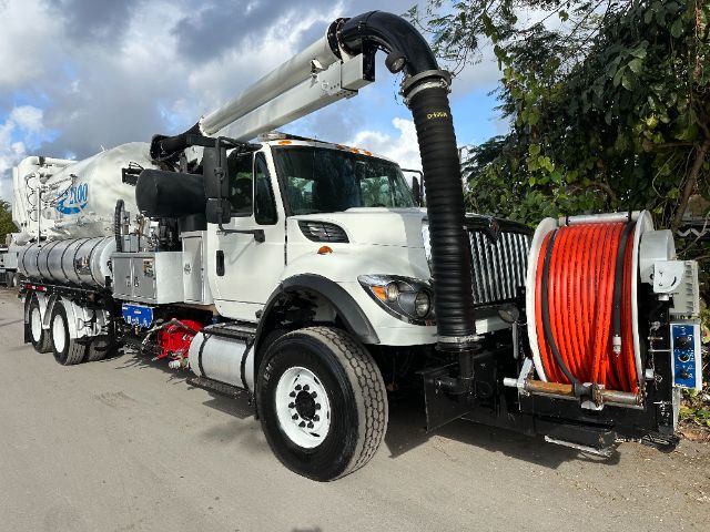 International 7500 vacuum truck with a large hose reel and rear vacuum unit, white cab, black intake hose, red hose on reel, side tanks, and industrial equipment.