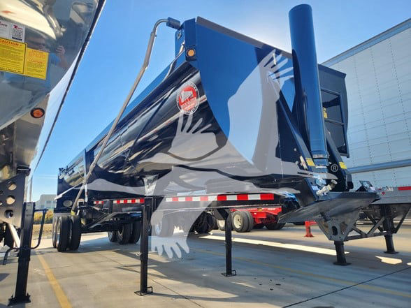 Trailer with blue/black paint, side view showing landing legs, hitch, and a blue vertical exhaust-like pipe; logo badge on side and reflective stripe along bottom edge.