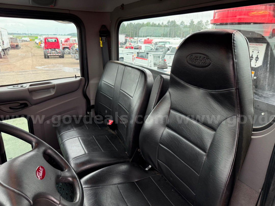 Peterbilt truck cab interior with black leather seats, steering wheel visible, window view of equipment yard.
