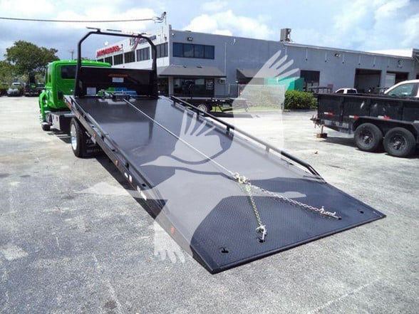 Tow truck flatbed with green cab, loading ramp extended in a parking lot, warehouse in background.