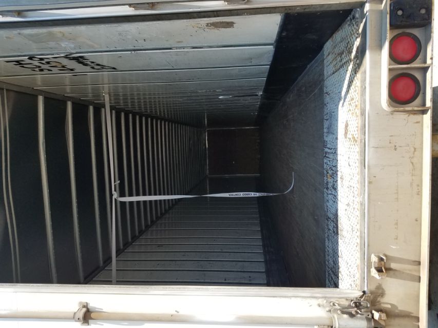 Trailer interior with metal walls, empty cargo space, strap hanging from ceiling, rear doors open, rubberized floor, some wear on the threshold.