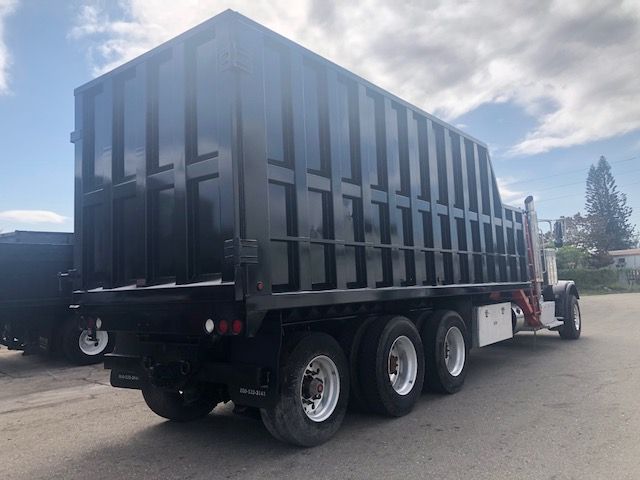 Black dump bed trailer attached to a Peterbilt truck with three rear axles, in a parking lot.