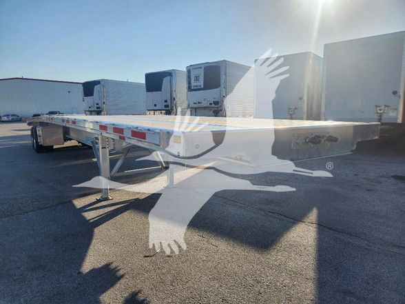 Flatbed trailer with long aluminum frame and landing gear, shown at a dealership lot with several trailer units in background.