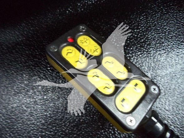 Truck part showing a compact black device with six yellow buttons/labels in a 2x3 arrangement. Each button has black symbols. A red indicator light is on the top left. The device is mounted on a black textured surface.