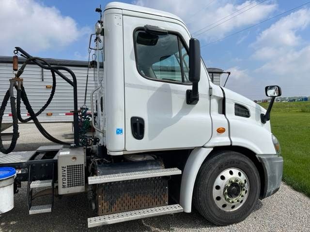 White Freightliner Cascadia cab with fuel tank, steps, and hydraulic lines visible. Side shows door with window, mirror, and wheel. Tank area labeled ‘HYDRIVE’ on rectangular component near steps.