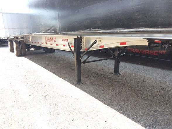 Flatbed trailer with a load covering tarp system and landing gear visible under the aluminum gooseneck. Side shows Reflective tape and Travers e height branding.
