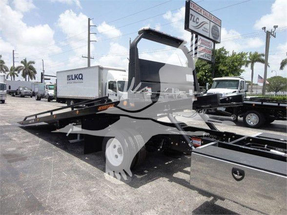 Isuzu NRR tow truck chassis with a flatbed; visible text includes TLC sign in background, FLKO logo on a white box truck, and no legible engine or fluid labels on the subject image.