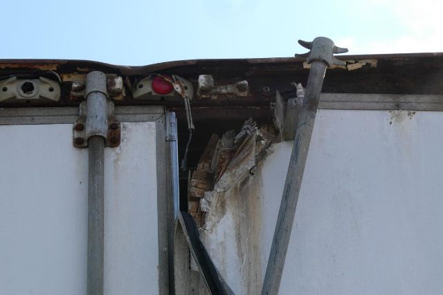 Rear door area of a trailer with a bent metal bar and damaged corner, showing hinges and a red linkage component.