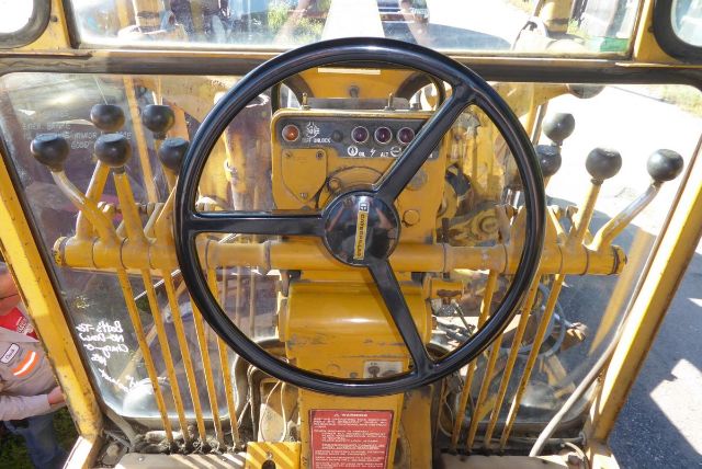 Caterpillar 16G grader operator cockpit with steering wheel, control levers on both sides, yellow body, dashboard with gauges.