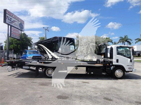 Isuzu NRR tow truck with flatbed, side view, white cab and black bed, parked at dealership lot with palm trees in background.
