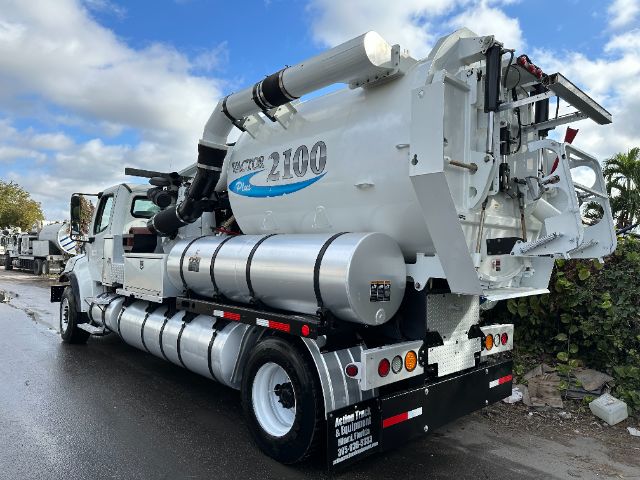Freightliner M2 vacuum truck with a white tank and rear hose assembly, side cylindrical tanks, and multiple hoses visible.