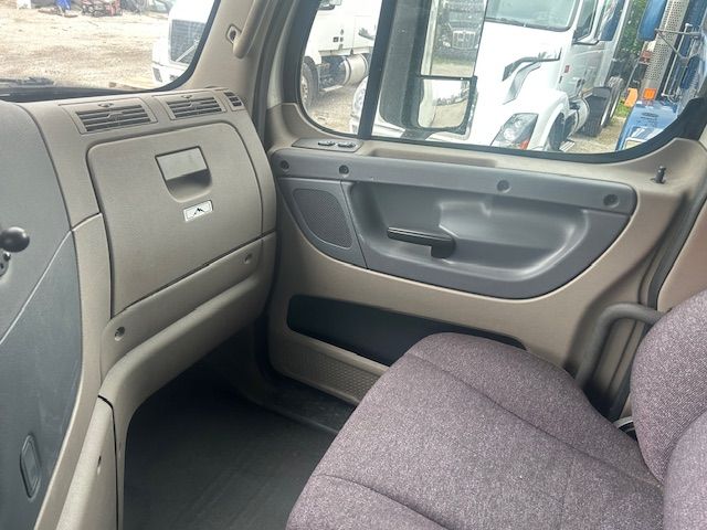 Truck interior: front passenger side dashboard with glove compartment, door panel, and passenger seat. Visible are the beige/gray plastic surfaces and fabric seat; windshield and side mirror reflections show other trucks in a yard.