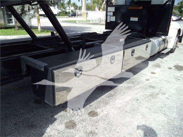 Kenworth T370 tow truck chassis with large rectangular tool/storage boxes along the side; some labels/stickers near the cab area; hydraulic components likely visible.