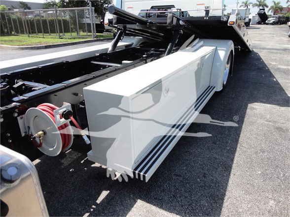 Truck chassis with hydraulic bumper/step and white storage box on side, red hose reel visible, flatbed with tilting mechanism in background.