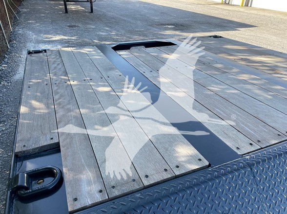 Trailer deck with wooden planks and a central slot, D-shaped tie-down ring visible near front edge, diamond plate edge. Includes watermark of birds in flight.