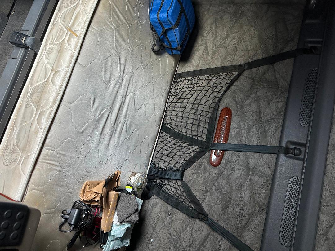 Truck cabin interior showing a bed with quilted mattress, netting storage on the wall, and various small items in a cluttered sleeper area.