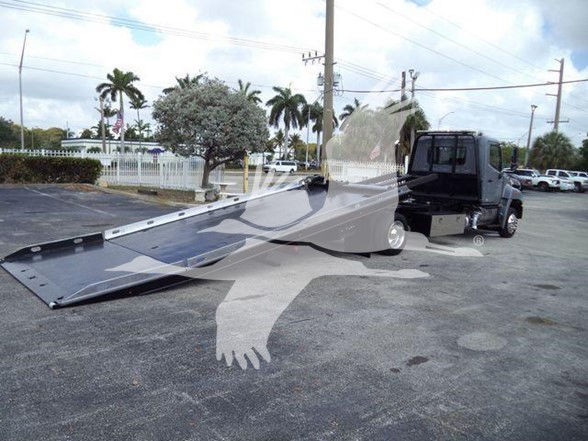 Tow truck with a long aluminum loading ramp extended from the rear of a black cab, in a parking lot.