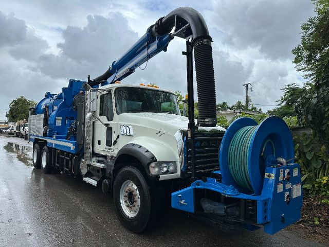 2017 Mack GU713 vacuum truck with long blue vacuum hose reel and pump system mounted on white truck cab, on a wet road.