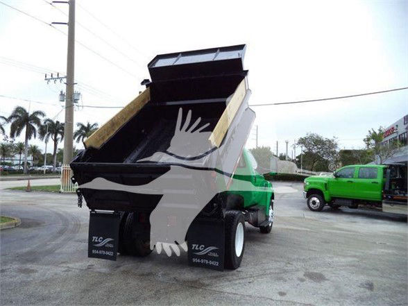 Dump truck in a dealership lot with its bed raised. Visible text: TLC ... 954-? numbers on the wheel well; no other legible labels clearly readable.