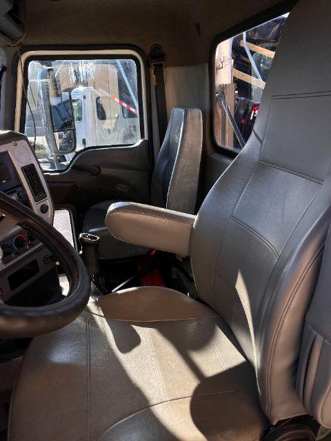 Truck cab interior from driver side showing steering wheel, seat, and passenger seat with armrest; dashboard and window view of another truck in yard.