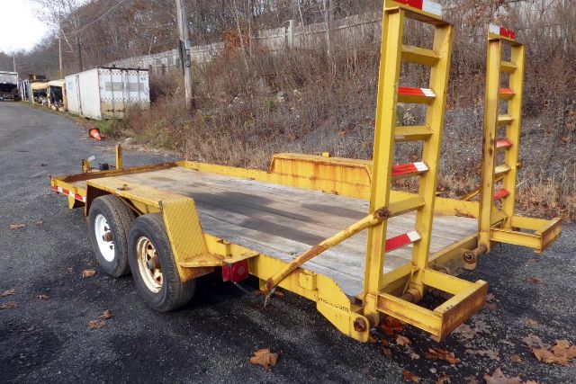 Trailer with dual-axle setup and ramp/swing-away loading ramps at rear, yellow painted steel frame, flatbed wooden deck, trailer hitch visible.