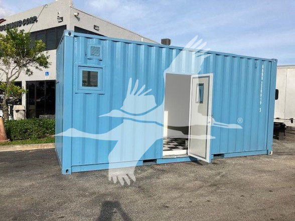 Blue shipping container trailer with a small window and an open door, sitting on a paved lot in front of a building.