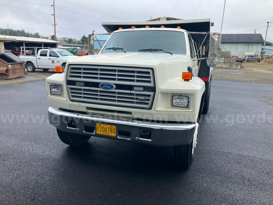 Front view of a white 1987 Ford F7000 dump truck with orange marker lights, diesel badge, Oregon plate E208780, and a black dump bed in the background.