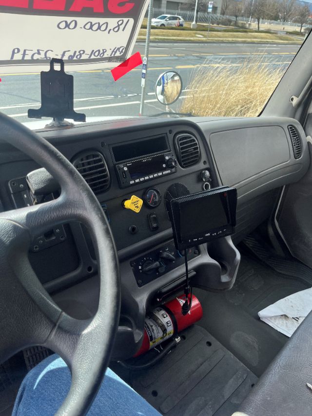 Cab interior of a Freightliner M2 106 box truck showing steering wheel, dashboard, radio, yellow hazard knob, tablet mount, fire extinguisher under center console, and a view through the passenger window.