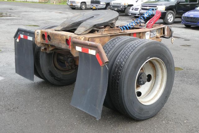 Trailer dolly with dual rear axles and large tires, rusty frame, spare tire mounted on top, mudflaps and red/blue coiled lines, Fruehauf EL FI model label visible.