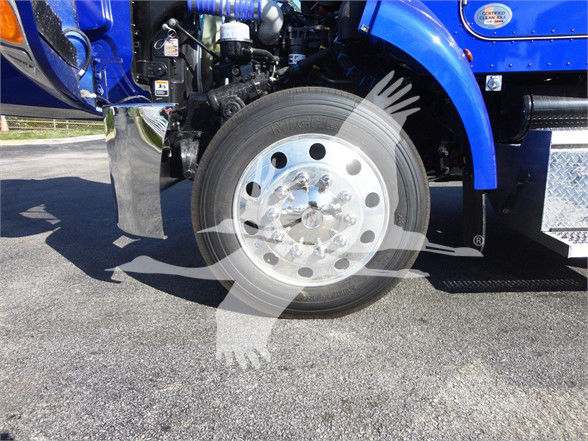Front wheel of a blue Peterbilt truck with chrome rim; engine bay area visible; circular watermark overlay.