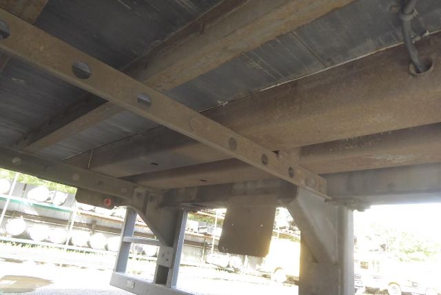 Trailer undercarriage showing steel I-beams, cross members, and suspension/frame components with a rusty finish and a tag hanging from the structure.