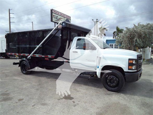 Truck showing a white Chevrolet Silverado 6500HD cab with a large black dump body. Visible text on the image includes a sign reading 'TRUCK SALES & SERVICE TLC' and the side of the truck may have a small badge but is not legible. The scene