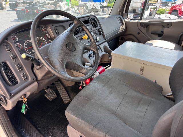 Truck interior showing steering wheel, dashboard, driver seat, and center console of a Freightliner M2 cab; large metal toolbox centered between seats; various controls on dash.