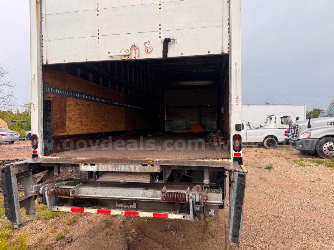 Truck cargo area of a 2016 Freightliner M2 106 box truck rear open, with loading lift at bumper, interior wooden paneling and empty cargo space.