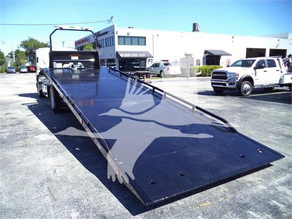 Isuzu tow truck flatbed with ramp extended in lot, showing black metal deck and rear section, D series cab in background.