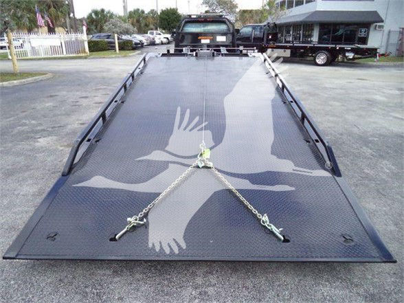 Flatbed tow truck trailer with a gray deck featuring a large white graphic hands silhouette chained in the center; chains and hooks visible on ramp; dealership lot in background.