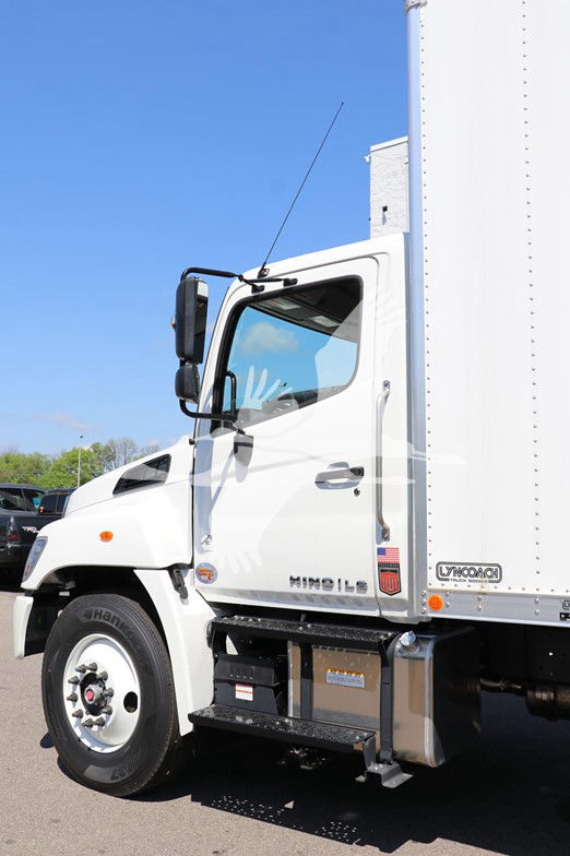Truck cab and box side showing Hino emblem, side steps, fuel tank, and front wheel; USA flag decal visible; white box truck with blue sky.