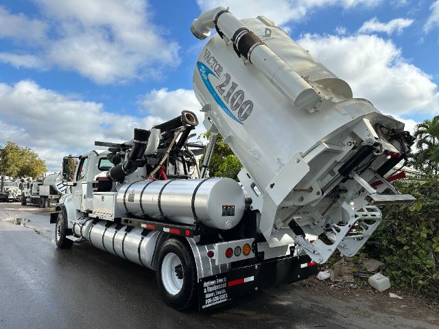 A Freightliner M2 vacuum truck with a large Vactor 2100 vacuum body raised at the rear. Side tanks, control panel, and hoses visible.