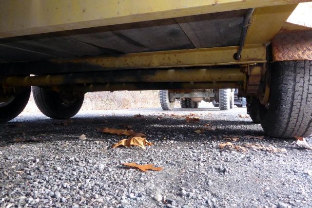 Trailer undercarriage showing frame rails and suspension with gravel ground and leaves, tires on both sides.
