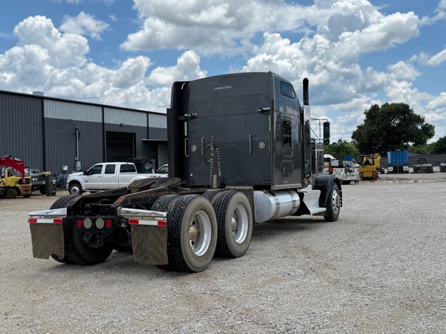 Kenworth W900 tractor in a used condition, black sleeper cab, triple rear axles, fuel tanks on both sides, gravel lot with industrial buildings.