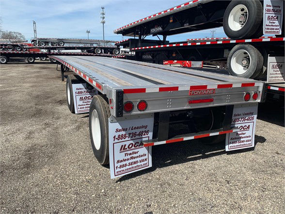Flatbed trailer with aluminum deck and dual rear axles, Fontaine logo on back, red taillights, stacked trailers in background, LOT signs on mudflaps.