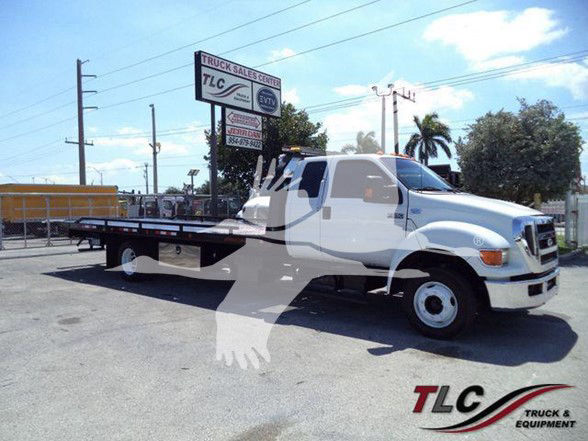 Tow truck with flatbed, Ford emblem on front, TLC Truck & Equipment watermark in lower right corner, dealership sign in background.