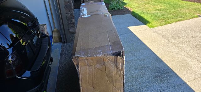 Boxed truck part or equipment in a residential driveway; long rectangular cardboard box on a concrete path next to a parked car.