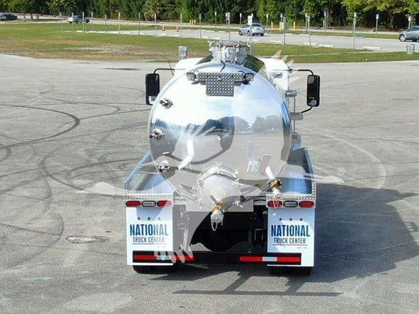 Rear view of a white 2023 Mack MD7 tanker truck equipped with a polished cylindrical stainless steel tank. Dual rear fenders with National Truck Center plates, taillights, and hoses visible on the back.