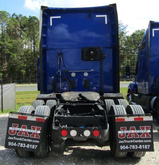 Rear view of a blue Kenworth T680 tractor with dual rear axles and mud flaps displaying Jax Truck Center branding and contact number.