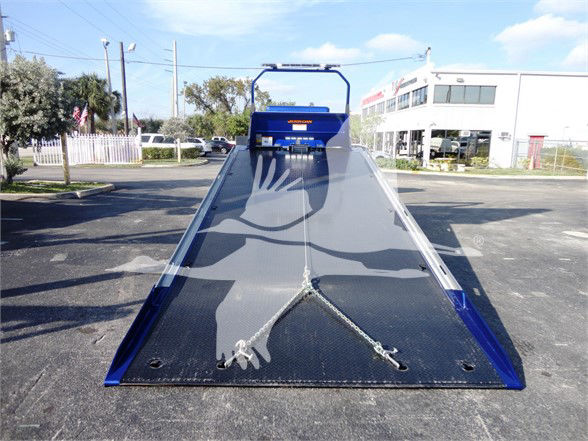 Tow truck flatbed with blue hydraulic equipment at the front; metal ramp extends toward camera with chains and hooks resting on surface.