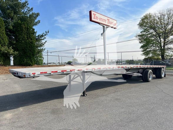 Flatbed trailer with dual rear axles and folded-down support leg, long steel deck, reflective red/white safety tape along sides, Idle stand and wheelset visible.